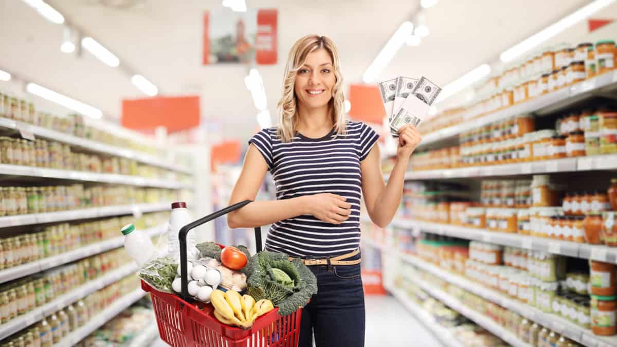 A woman standing in a grocery store aisle holding a basket with food and money.