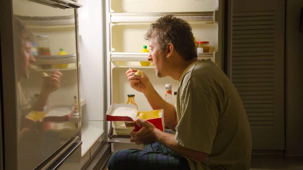A man eating in front of an open fridge at night.