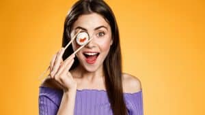 A girl holding a piece of sushi in front of her eye.