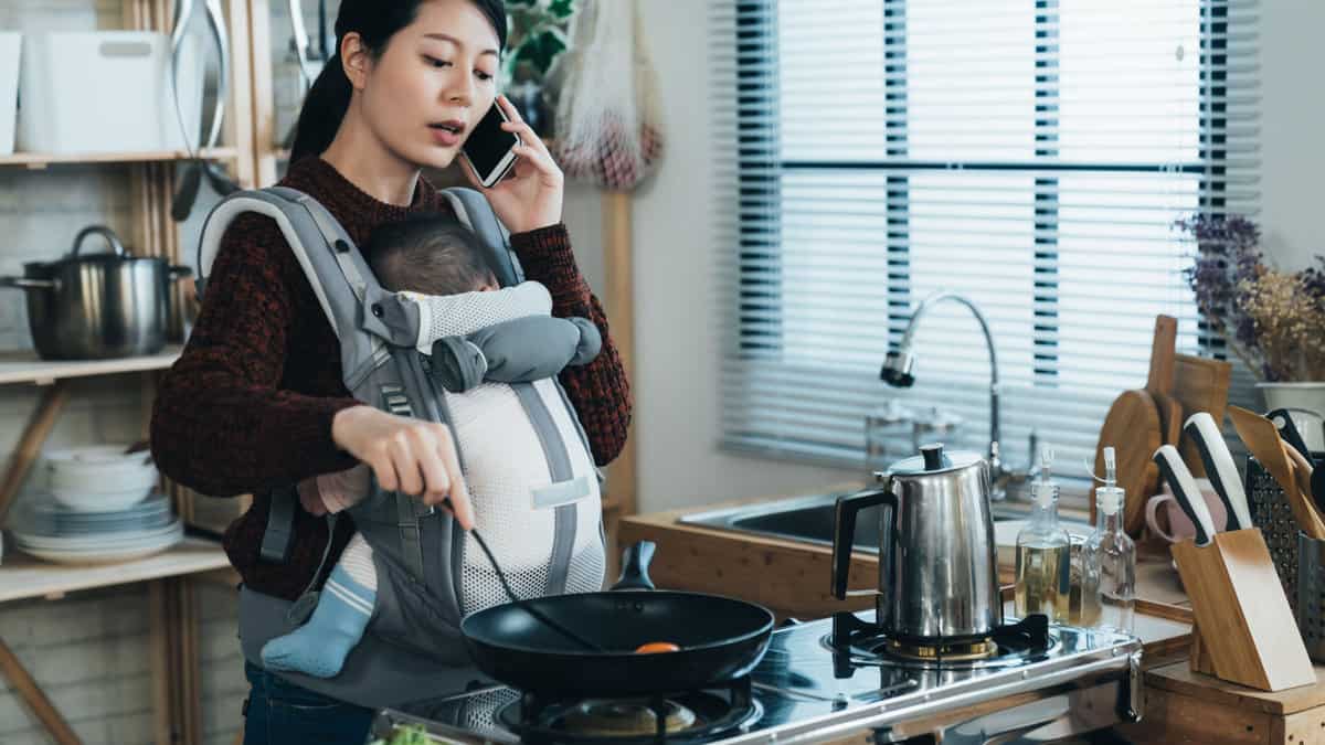A pregnant woman cooking with a child in a carrier and talking on the phone.