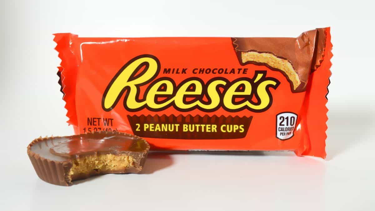 A partially eaten Reese's peanut butter cup in front of a package of peanut butter cups with a white background.