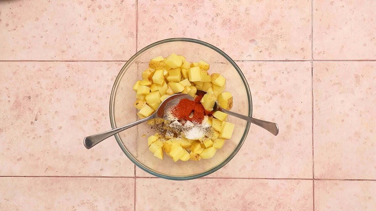 Potato cubes in a bowl topped with paprika powder, garlic powder, salt, pepper and sugar, before mixing.