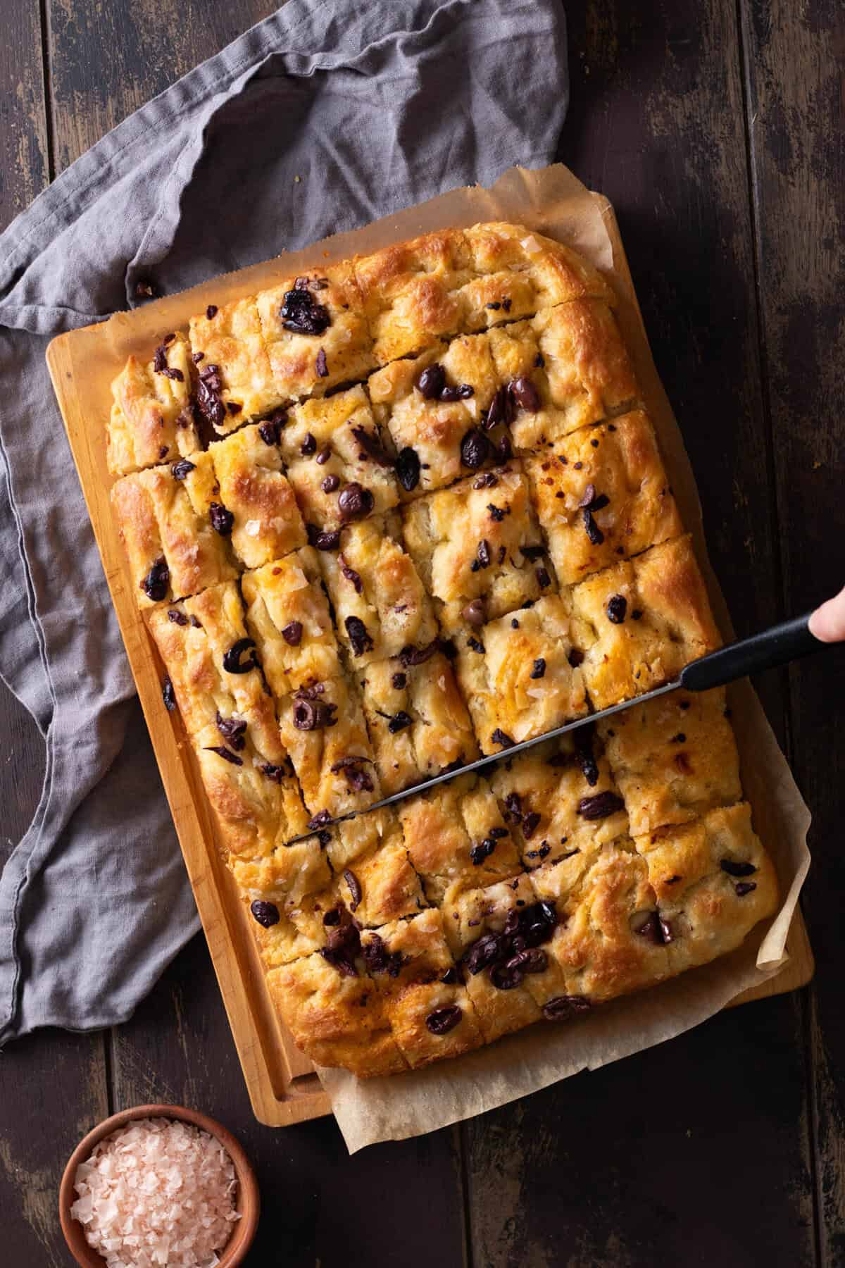 Top down view of an easy overnight focaccia topped with black olives, showing a knife cutting it into squares.