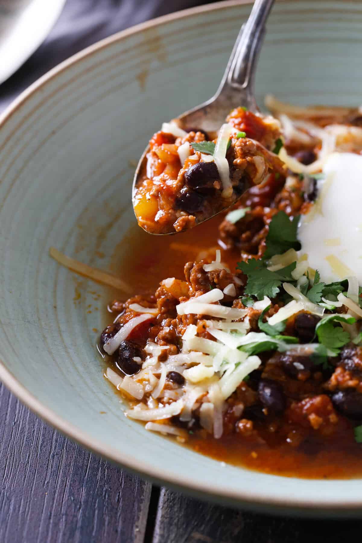 A green bowl with beef chili topped with sour cream, shredded cheese and fresh cilantro.