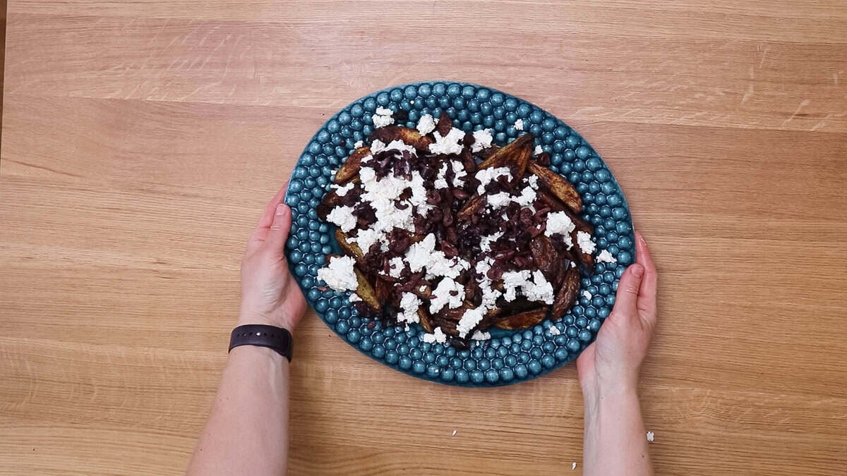 Baked crispy potato wedges on a blue plate, topped with feta cheese and black olives.